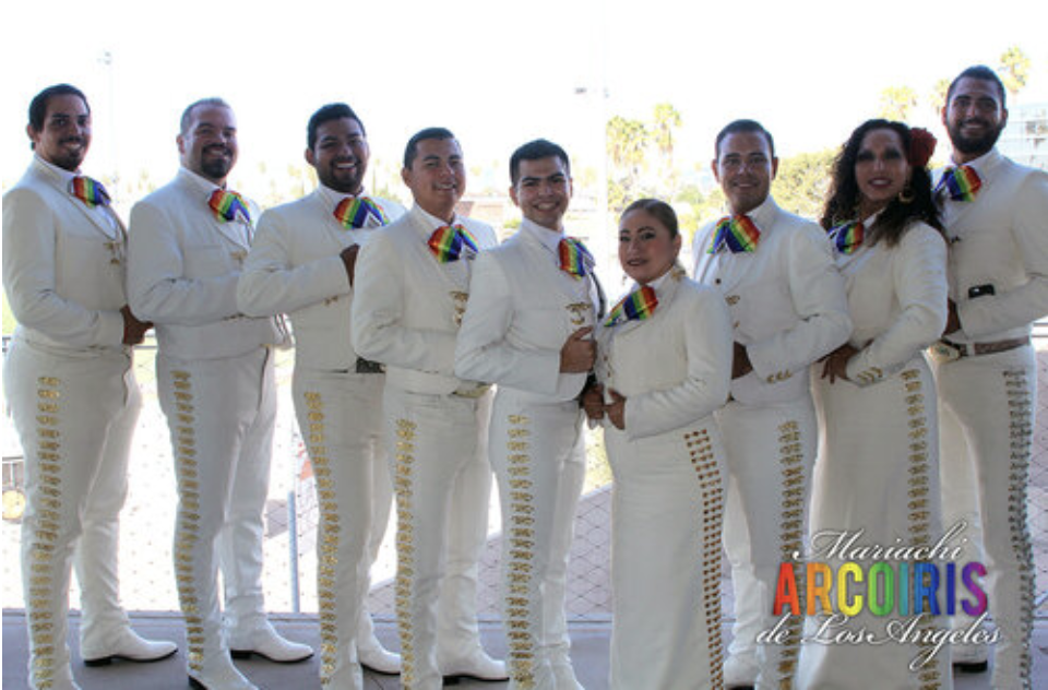 Photo of Mariachi Arcoiris de Los Angeles. There are nine mariachi performers wearing white outfits and rainbow colored ties